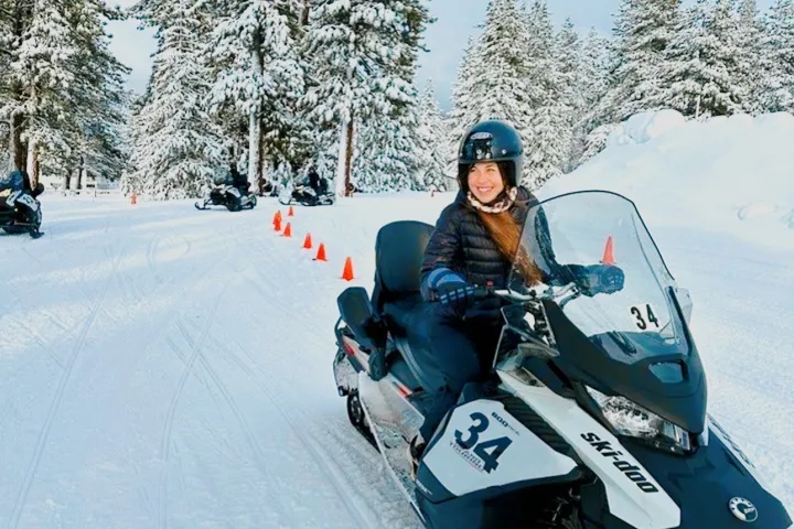 Person on snowmobile with helmet, snowy trees and cones in background.