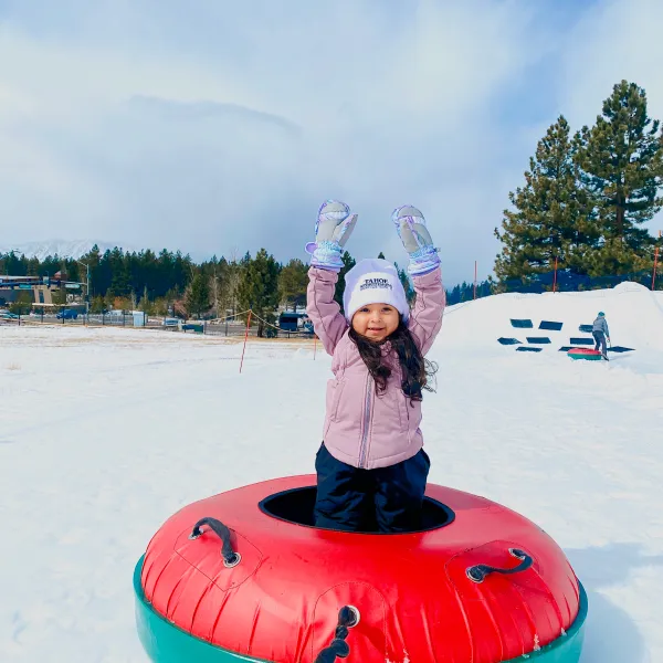 Child in snow wearing winter clothes, standing in a colorful inflatable tube with trees and sky in the background.