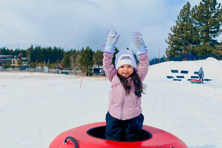 Child in snow wearing winter clothes, standing in a colorful inflatable tube with trees and sky in the background.