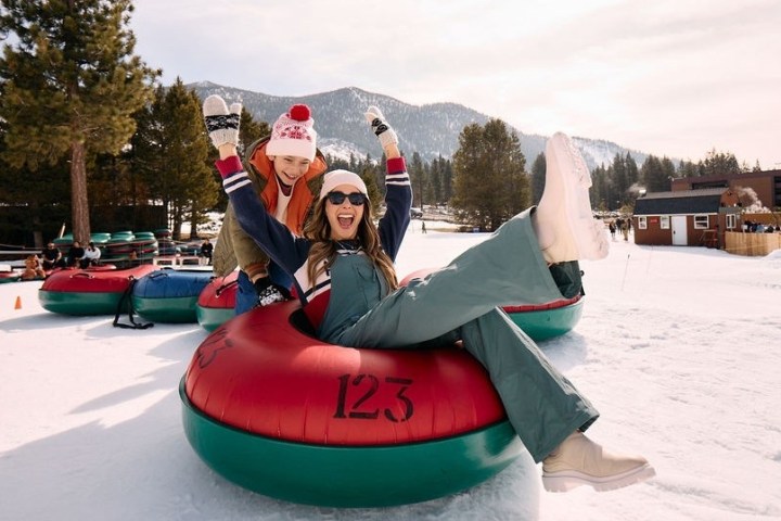Two people enjoying snow tubing, laughing and raising hands, with mountains and trees in the background.